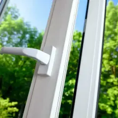 Uchylne okno z białą klamką, widok na zielone drzewa. Dowiedz się, jak zdjąć skrzydło z okna uchylnego.