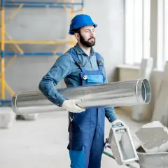 Budowlaniec w niebieskim kasku i kombinezonie niesie metalową rurę, przygotowując się do prac związanych z tym, jak ocieplić rury od rekuperacji.