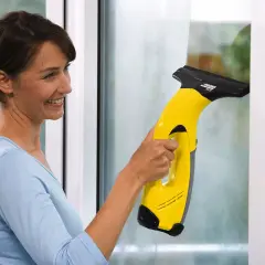 Kobieta z uśmiechem czyści okno żółtym odkurzaczem Karcher. Dowiedz się, dlaczego zostają smugi po myjce Karcher i jak ich unikać.