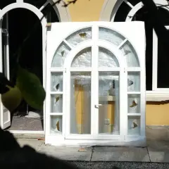 Stare okna z pękniętymi szybami czekają na wywóz. W tle widać otwarte drzwi i wnętrze budynku.