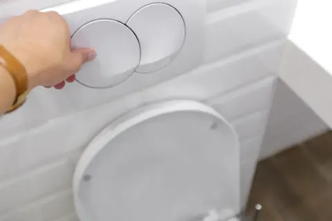 Dłoń naciska na okrągły guzik do spłuczki nad sedesem.