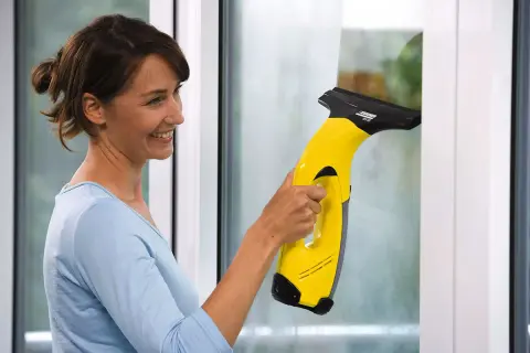 Kobieta z uśmiechem czyści okno żółtym odkurzaczem Karcher. Dowiedz się, dlaczego zostają smugi po myjce Karcher i jak ich unikać.