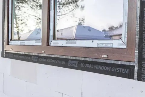 Montaż okna w białym pustaku z brązową ramą, widoczna taśma Soudal Window System. Dowiedz się, ile kosztuje montaż okna.