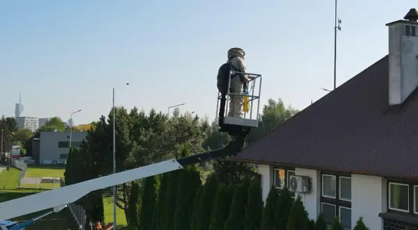 Osoba w stroju pszczelarza na podnośniku koszowym przy dachu domu, przygotowuje się do akcji jak odstraszyć osy z poddasza.
