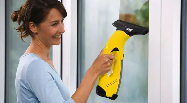 Kobieta z uśmiechem czyści okno żółtym odkurzaczem Karcher. Dowiedz się, dlaczego zostają smugi po myjce Karcher i jak ich unikać.